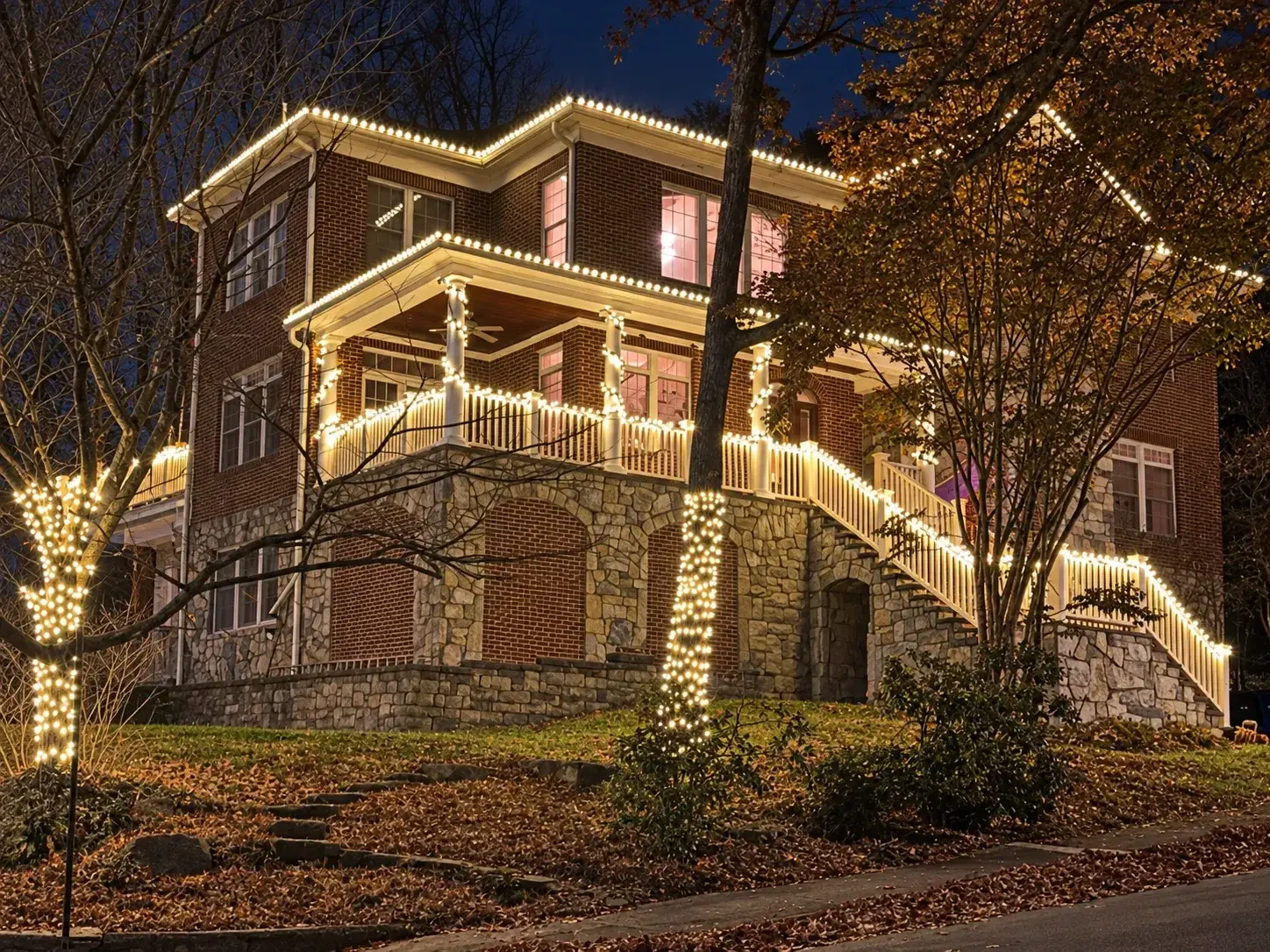 A three-story brick and stone house on a hill lit by Blink: warm white C9s on every roofline, the porch columns and railings, the long side staircase, and two wrapped tree trunks.