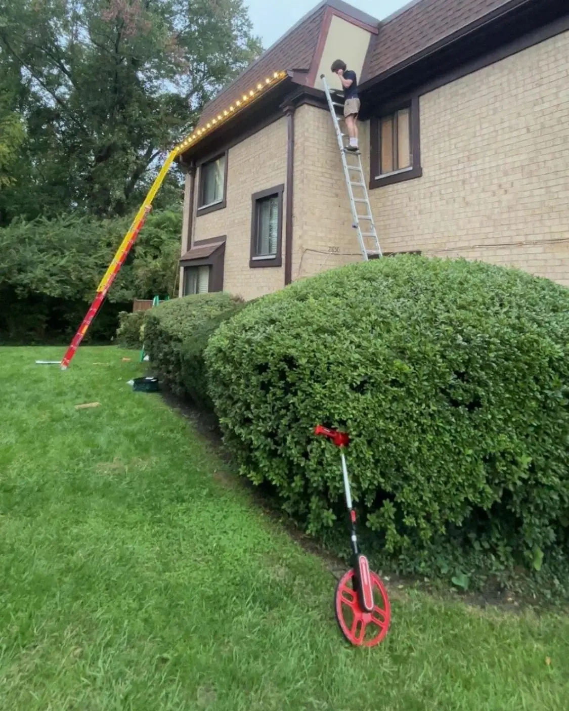 A Blink installer on a ladder clipping warm white C9 lights along the gutter of a two-story brick house, with a second ladder against the finished run and a measuring wheel on the lawn.