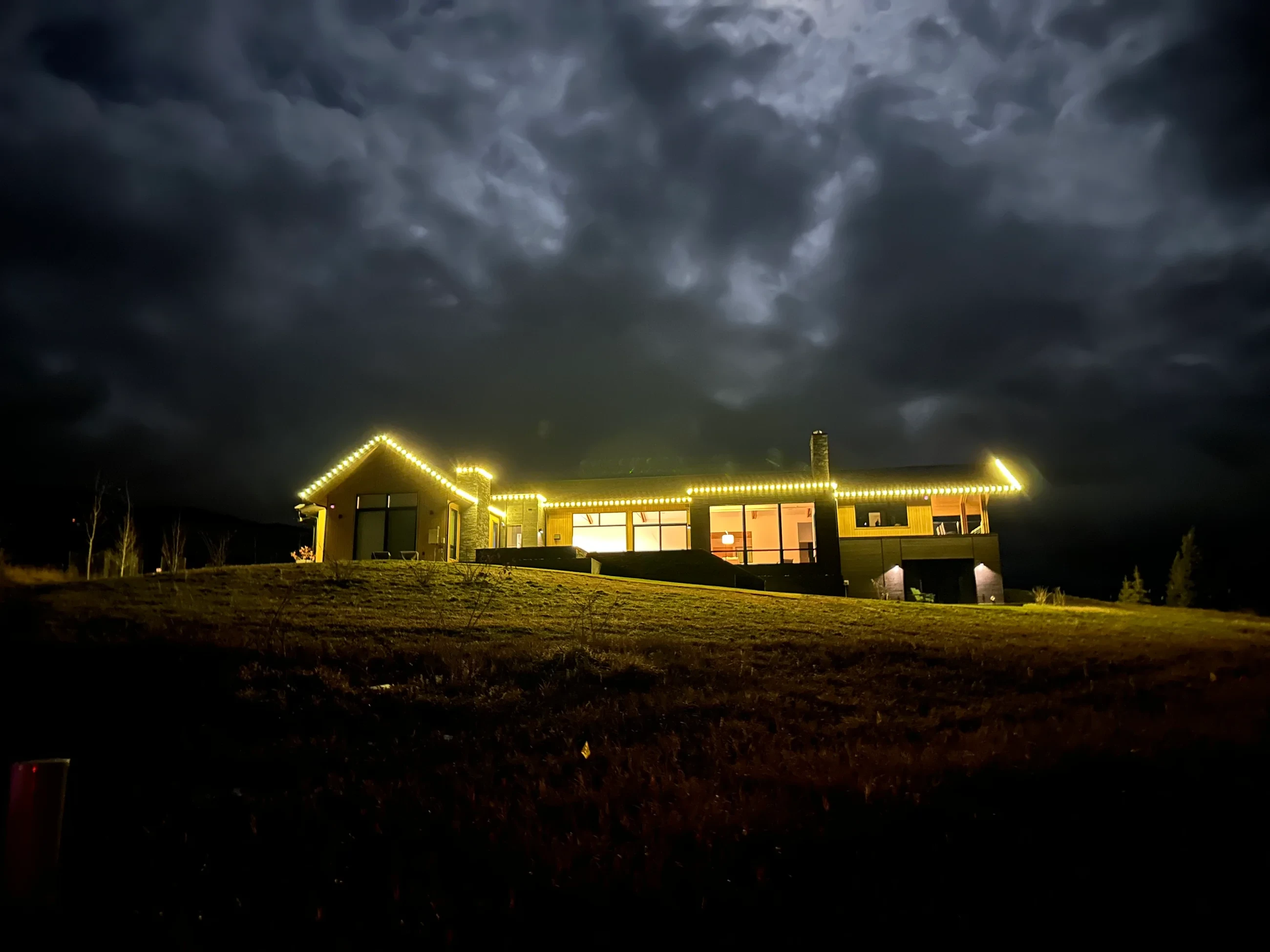 Single-story home outlined in gold roofline lights beneath dark storm clouds.