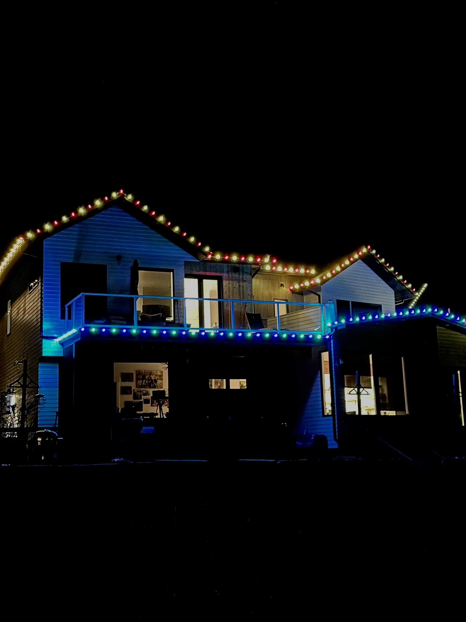 Two-story home with red and warm white upper rooflines and blue and green deck lights.