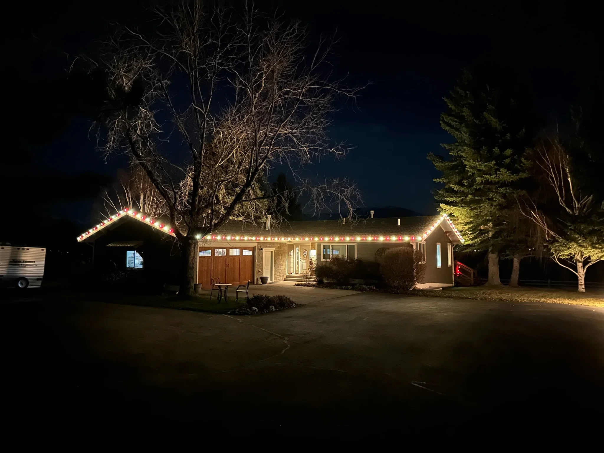 Ranch-style home with alternating red and white roofline lights under tall trees.