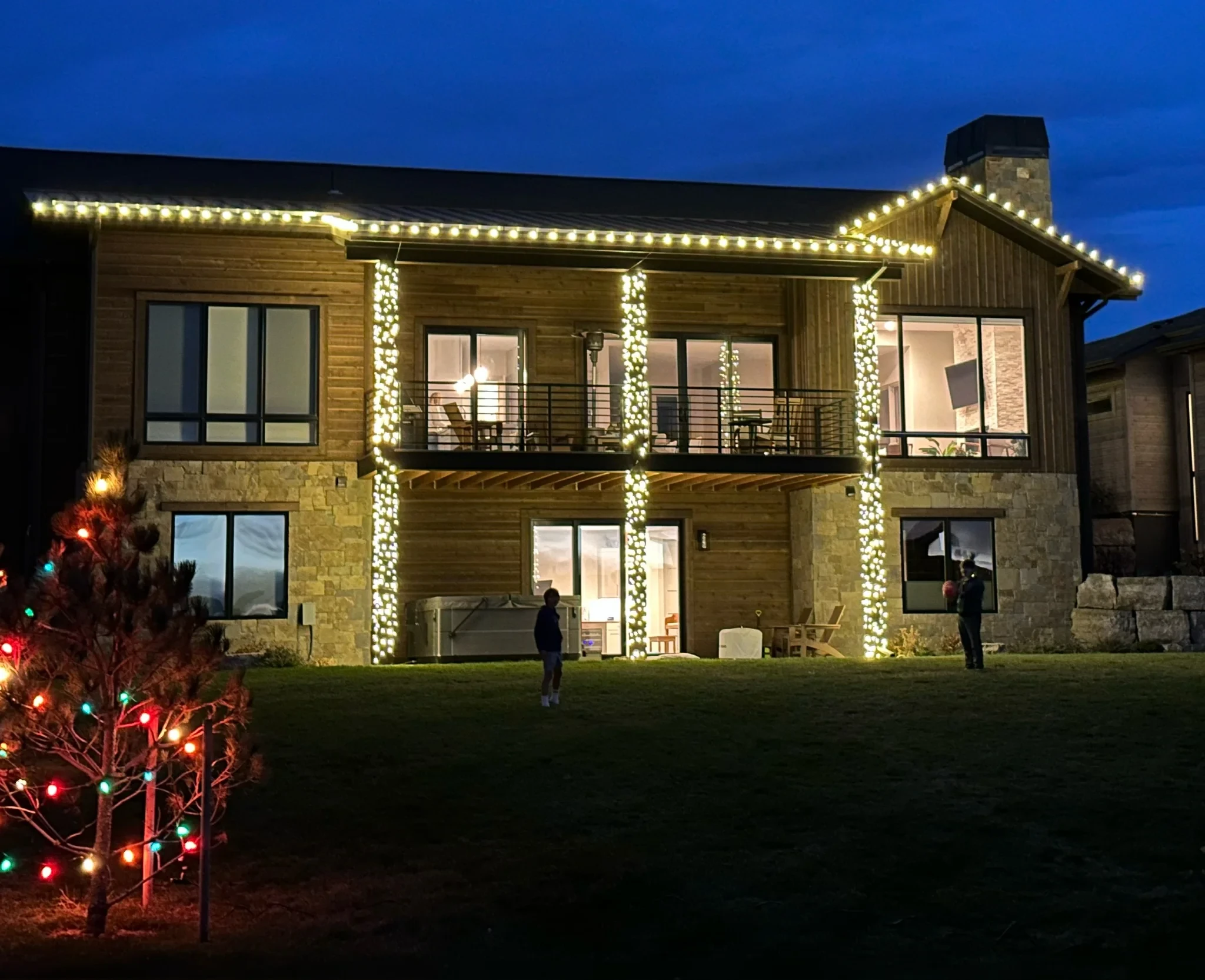 Two-story stone and timber home outlined in warm white roofline lights, with wrapped porch pillars.