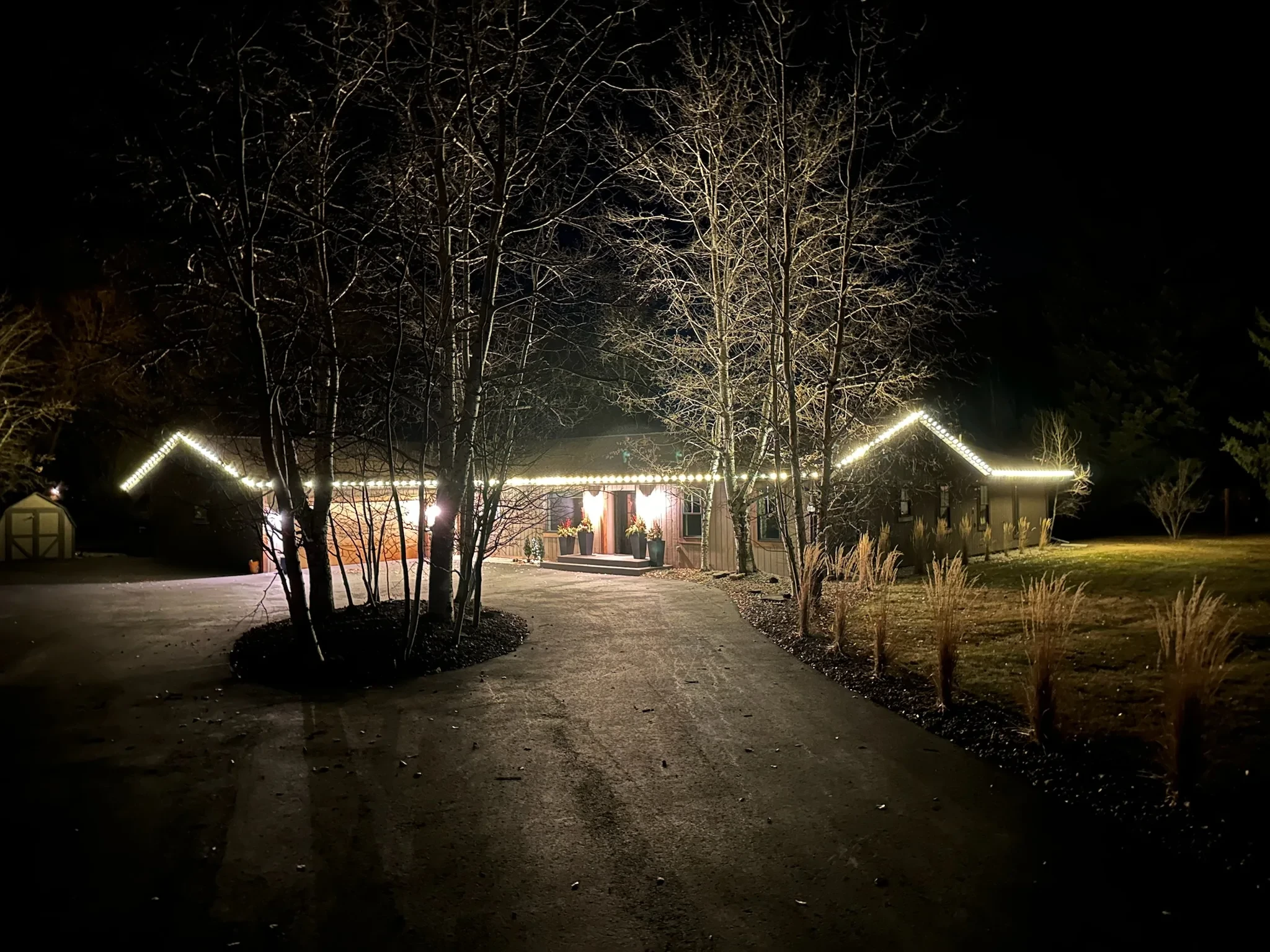 Single-story home with warm white roofline lights, seen down a long driveway through bare trees.