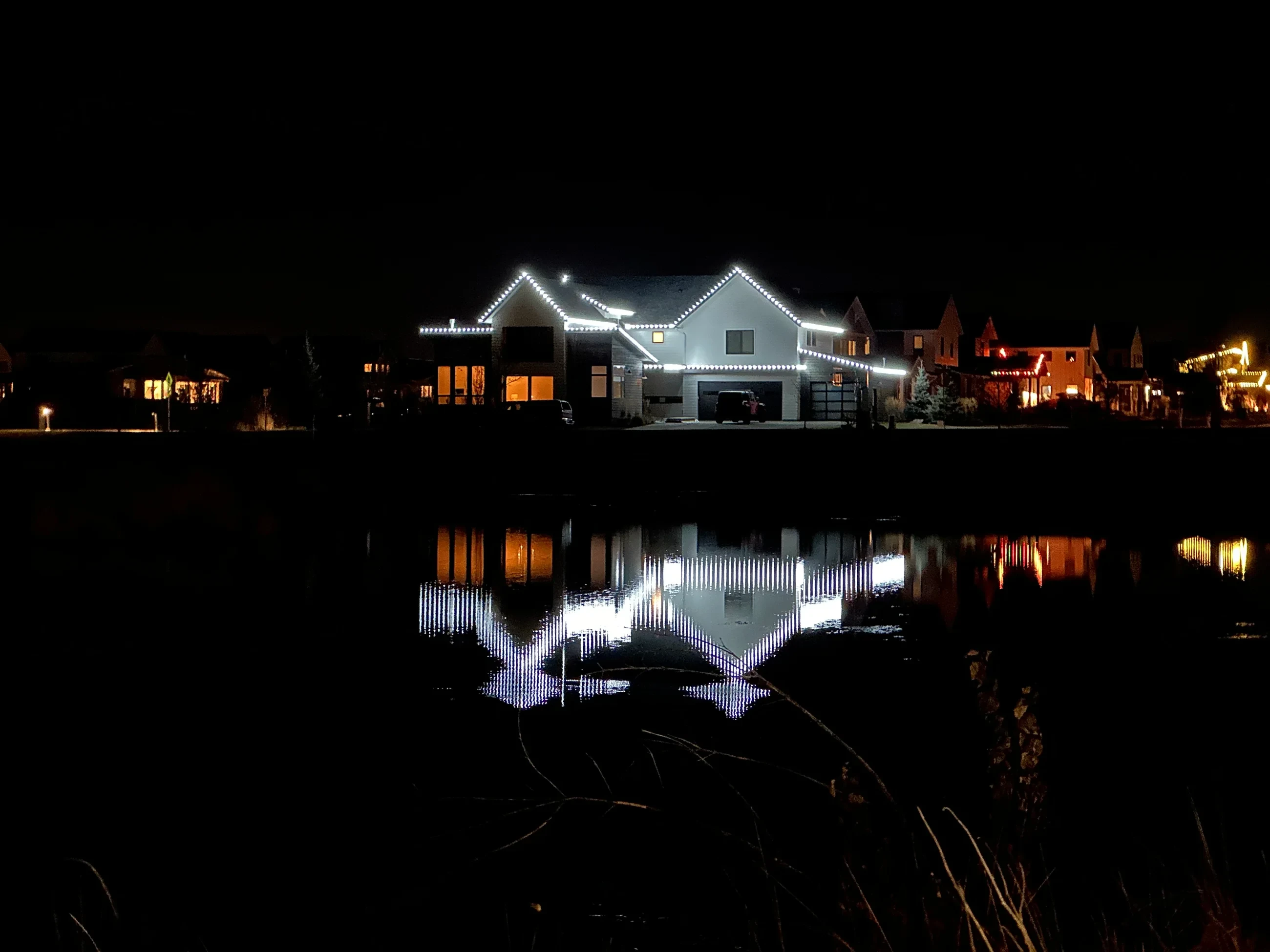 Home outlined in white roofline lights, reflected in a still pond at night.